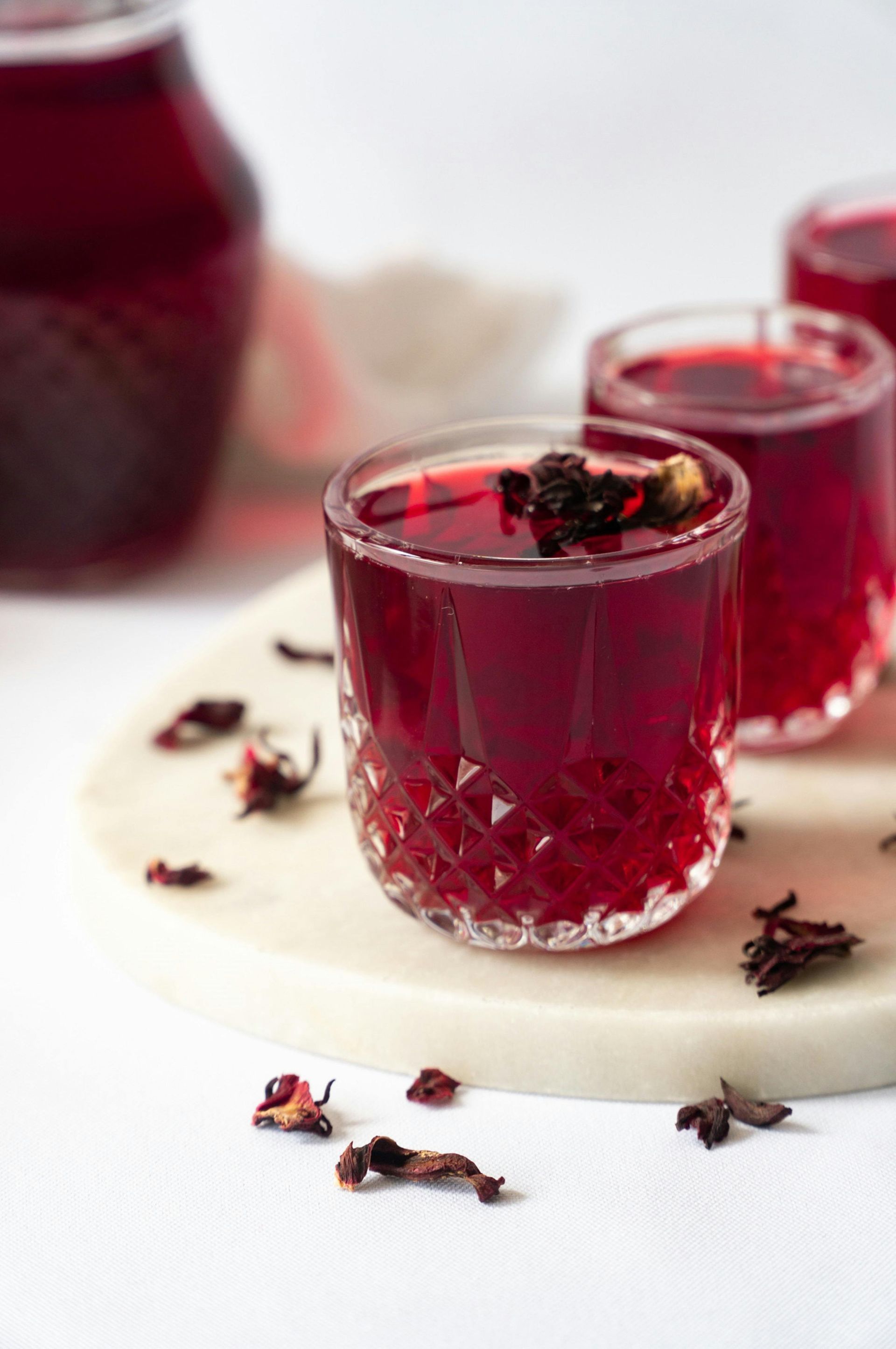 Deep ruby red hibiscus tea served in faceted glass cups, garnished with dried flower petals on a white marble board.