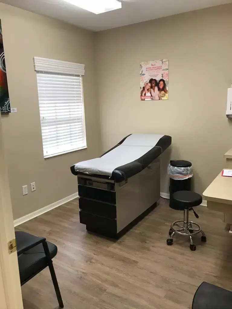 Examination room with a patient bed, a stool, and a small desk; beige walls and wood-look flooring.