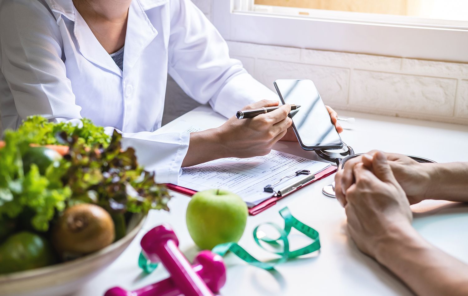 Dietitian pointing to a phone screen, discussing diet with a patient. Fresh produce and fitness equipment are on the table.