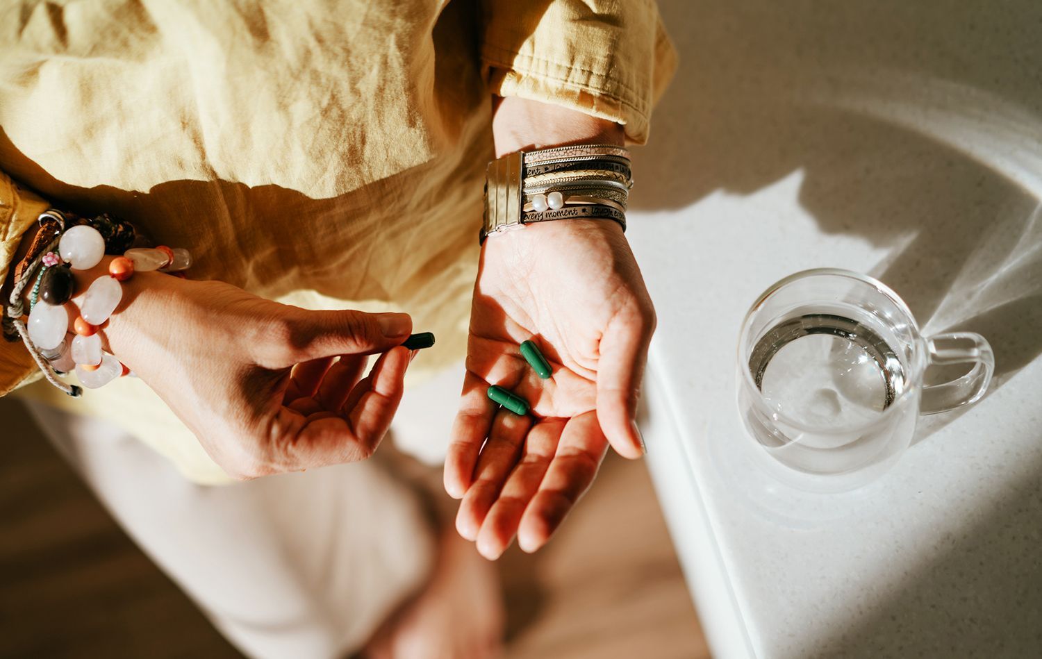 Person holding pills, another hand near a glass of water.