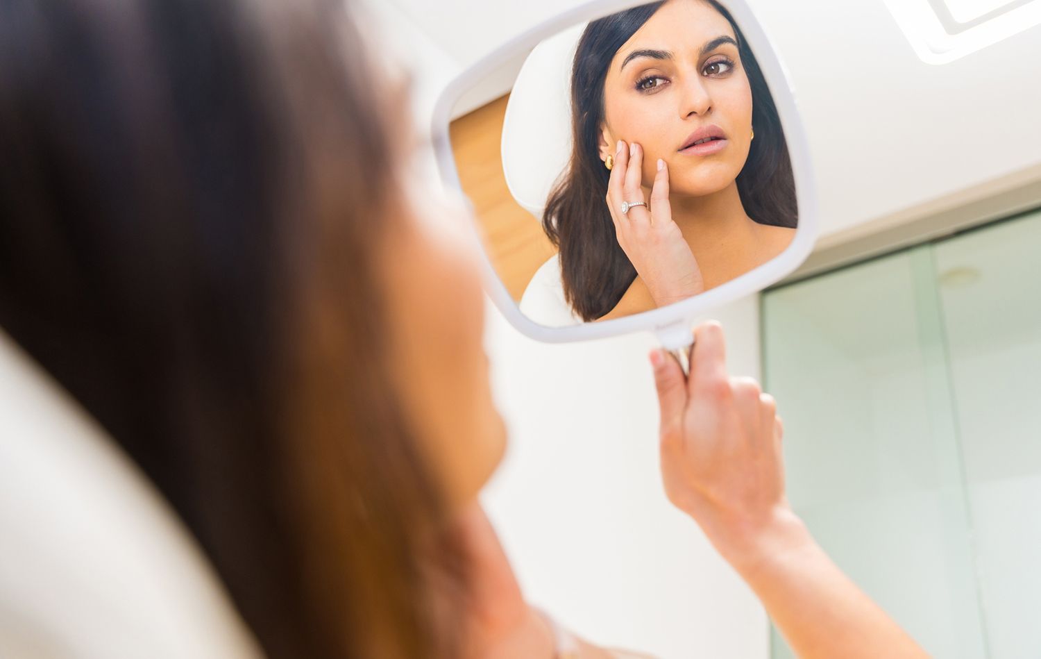 Woman examines her face in a handheld mirror, lit in a bright room. Woman examines her face in a handheld mirror, lit in a bright room.