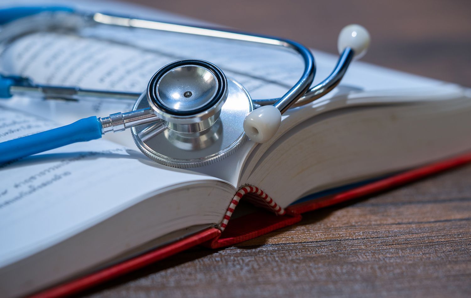 Blue stethoscope rests on an open book with red cover on wooden surface. Blue stethoscope rests on an open book with red cover on wooden surface.