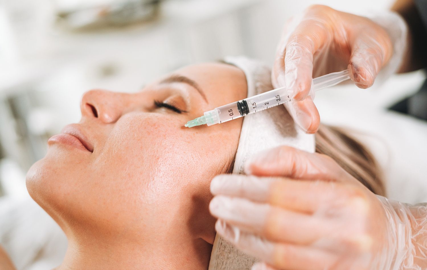 A woman receives a cosmetic injection near her eye, administered by a gloved hand in a medical setting. A woman receives a cosmetic injection near her eye, administered by a gloved hand in a medical setting.
