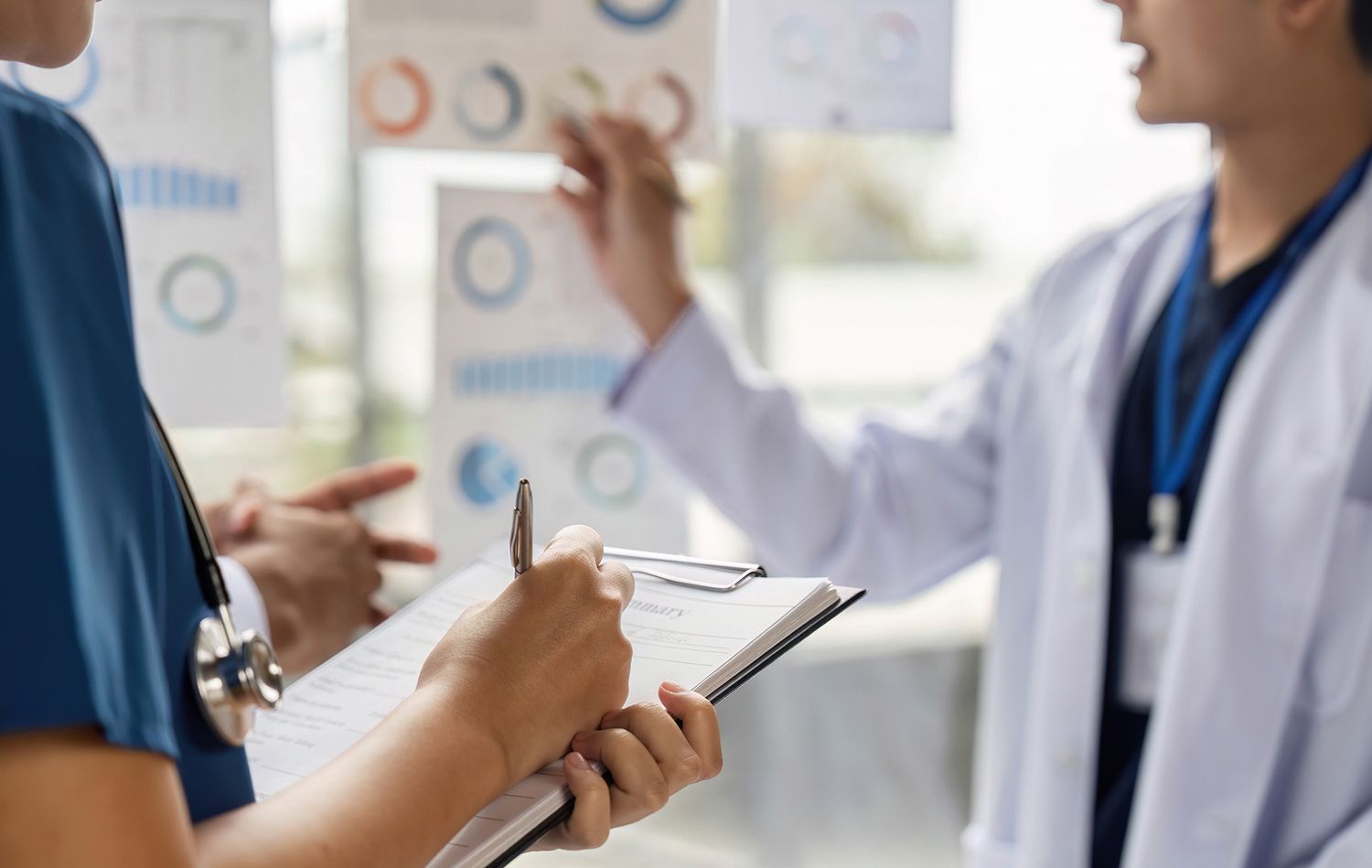 Two medical professionals analyzing charts, one writing on a clipboard, in a bright office. Two medical professionals analyzing charts, one writing on a clipboard, in a bright office.