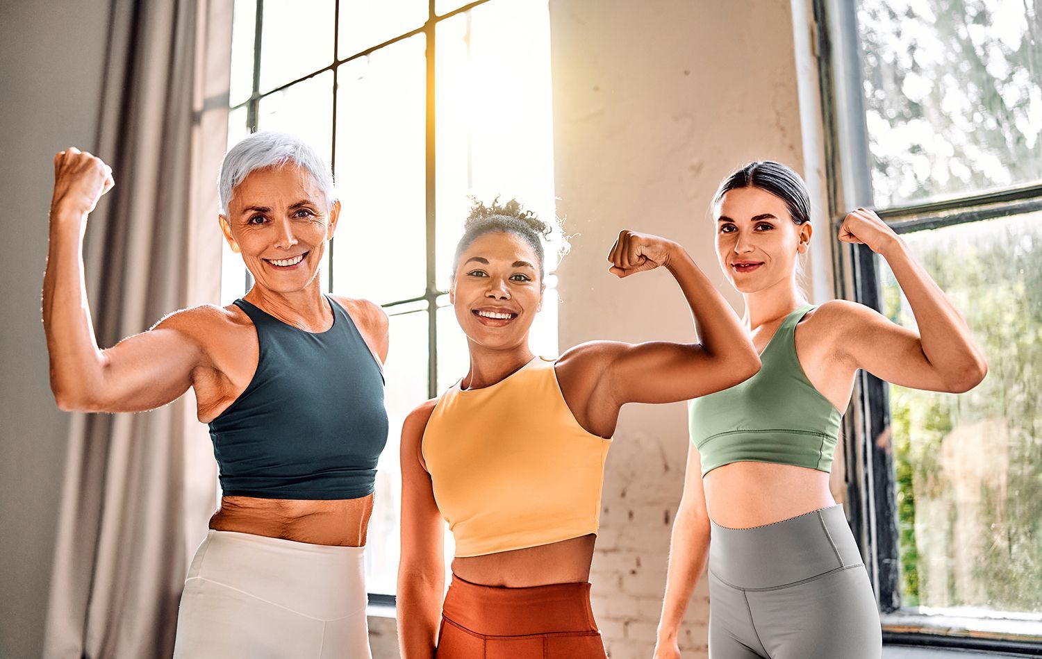 Three women in workout clothes flexing arms, smiling, in a sunlit room with windows. Three women in workout clothes flexing arms, smiling, in a sunlit room with windows.