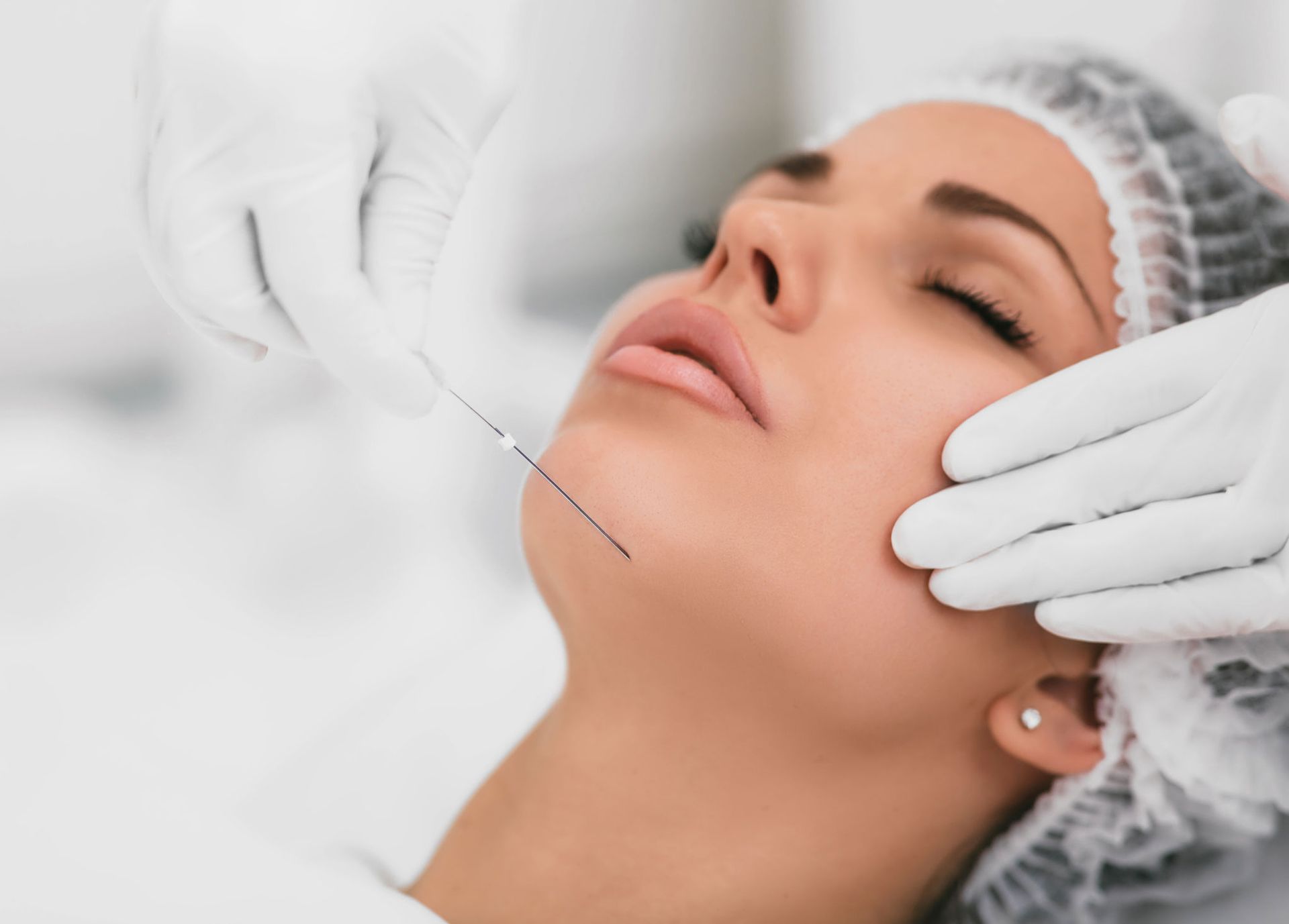 Woman receiving a cosmetic procedure on her chin; doctor's gloved hands, needle, and white setting. Woman receiving a cosmetic procedure on her chin; doctor's gloved hands, needle, and white setting.
