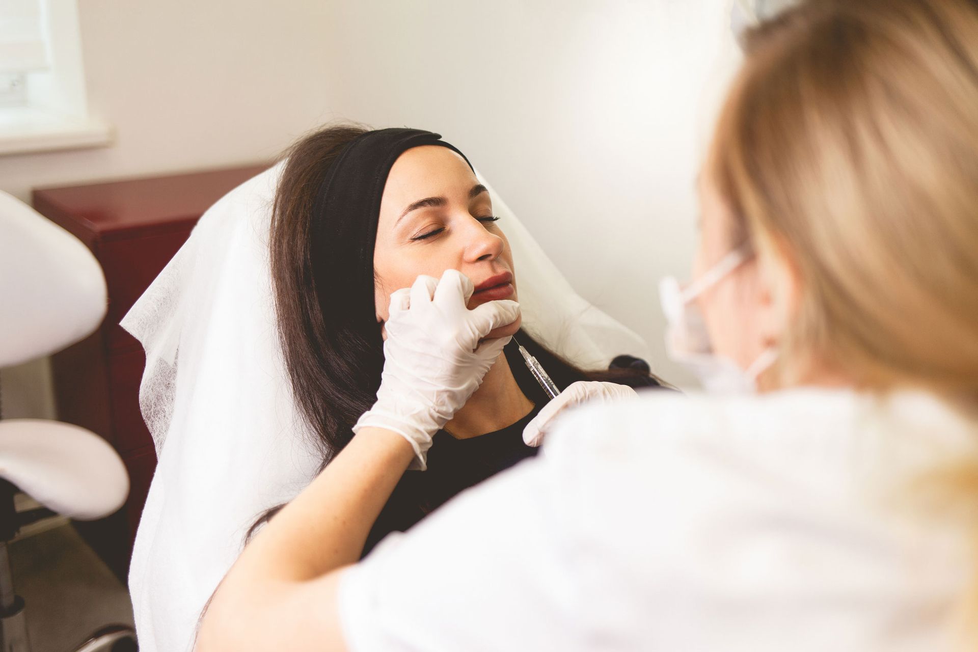 Person receiving lip injections from a person wearing gloves and a mask in a medical setting. Person receiving lip injections from a person wearing gloves and a mask in a medical setting.
