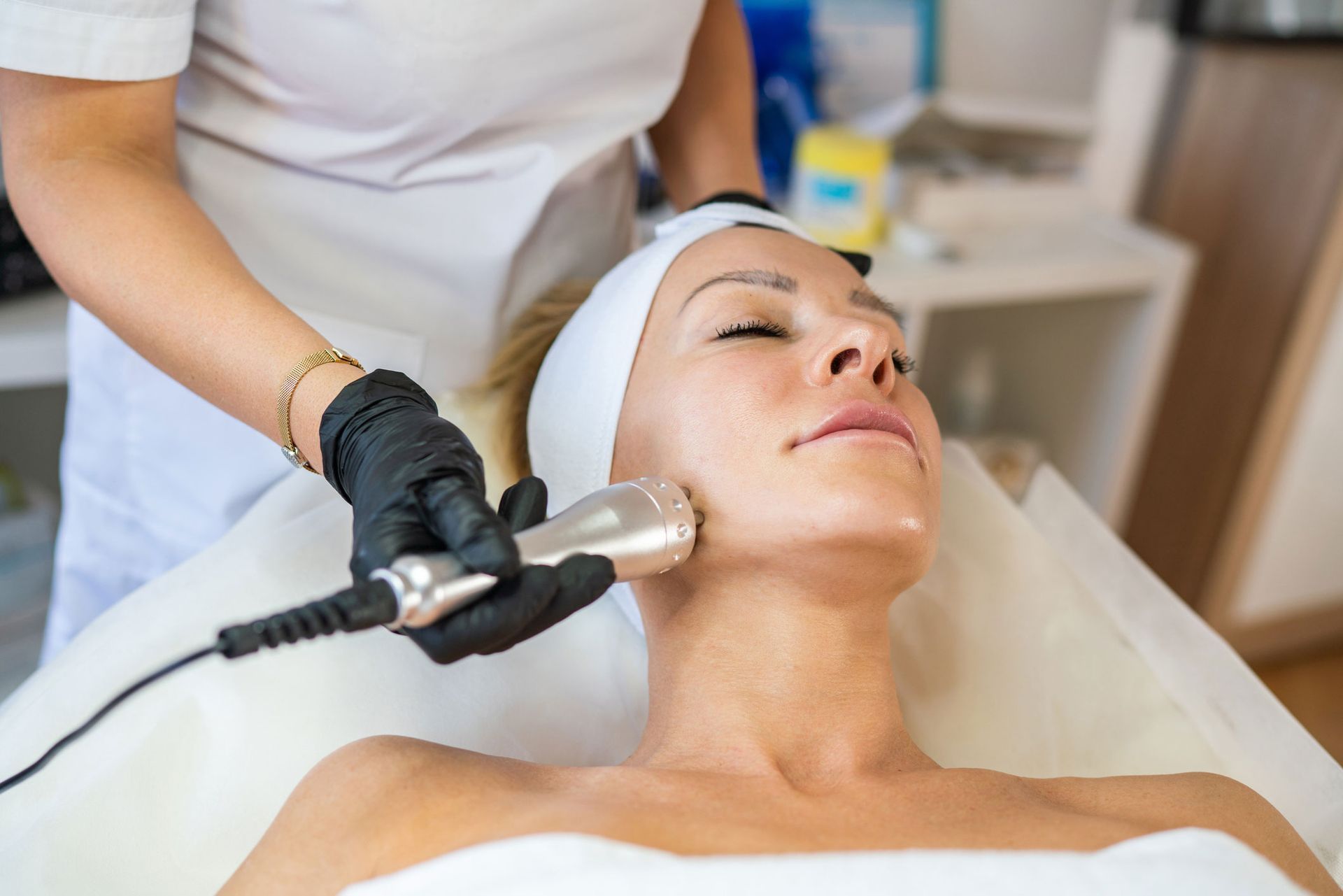 Woman receiving facial treatment with a handheld device. Therapist in black gloves in a spa setting.
