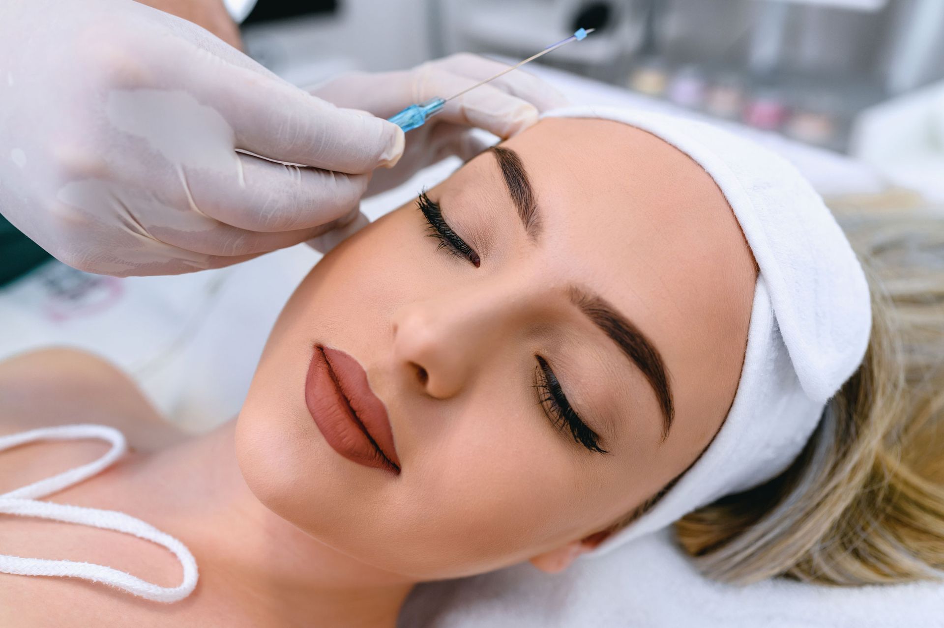 Person receiving a cosmetic procedure on the forehead. Hands with gloves hold a needle, and the person has eyes closed. Person receiving a cosmetic procedure on the forehead. Hands with gloves hold a needle, and the person has eyes closed.