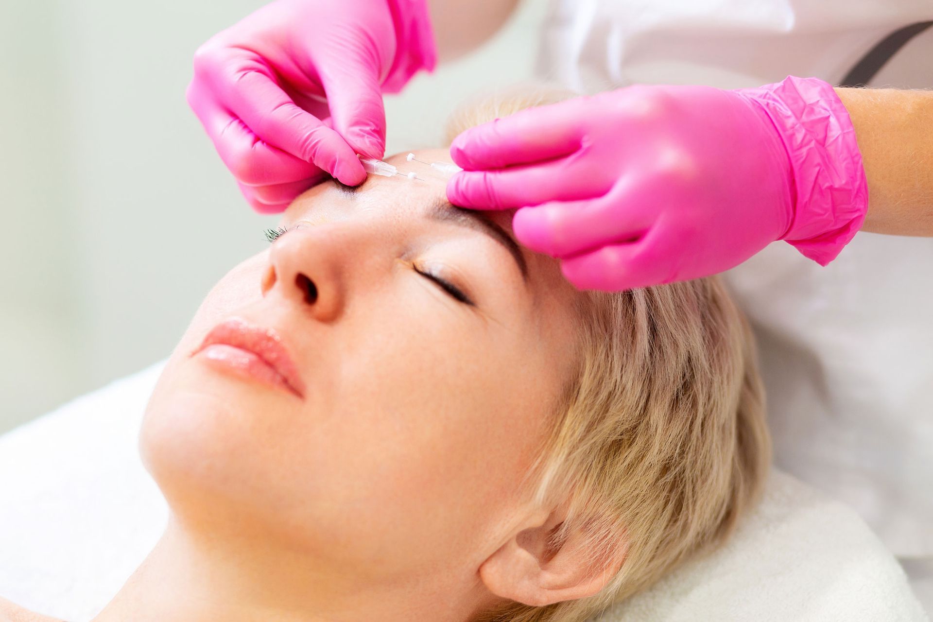 Woman receiving facial treatment, pink-gloved hands applying product to her forehead, eyes closed. Woman receiving facial treatment, pink-gloved hands applying product to her forehead, eyes closed.
