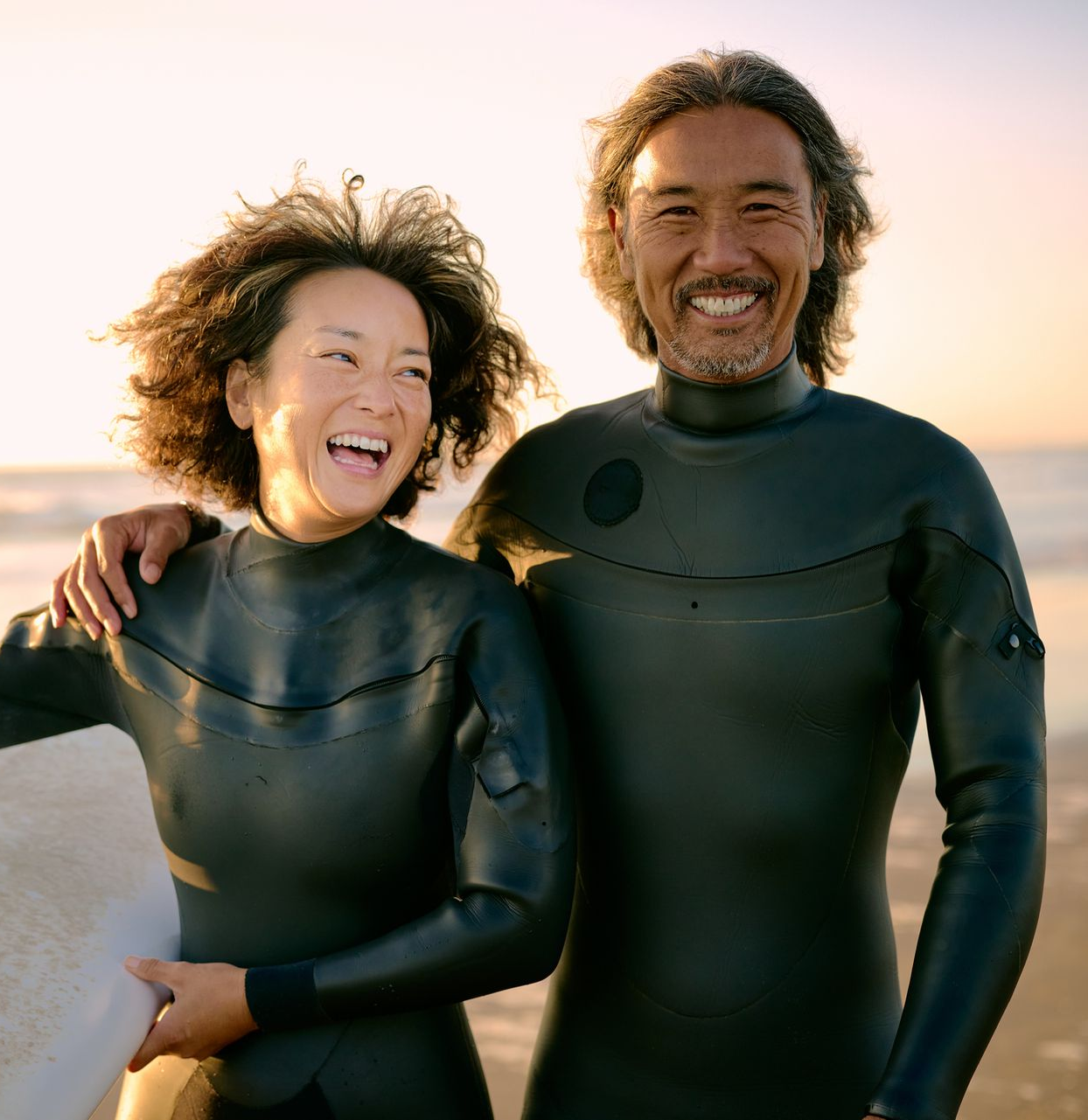Couple in wetsuits on beach, holding surfboard, smiling.