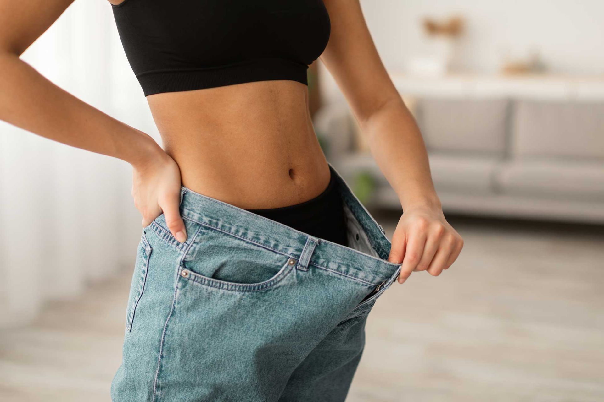 Woman wearing loose jeans, showing weight loss; standing indoors with hand on hip.
