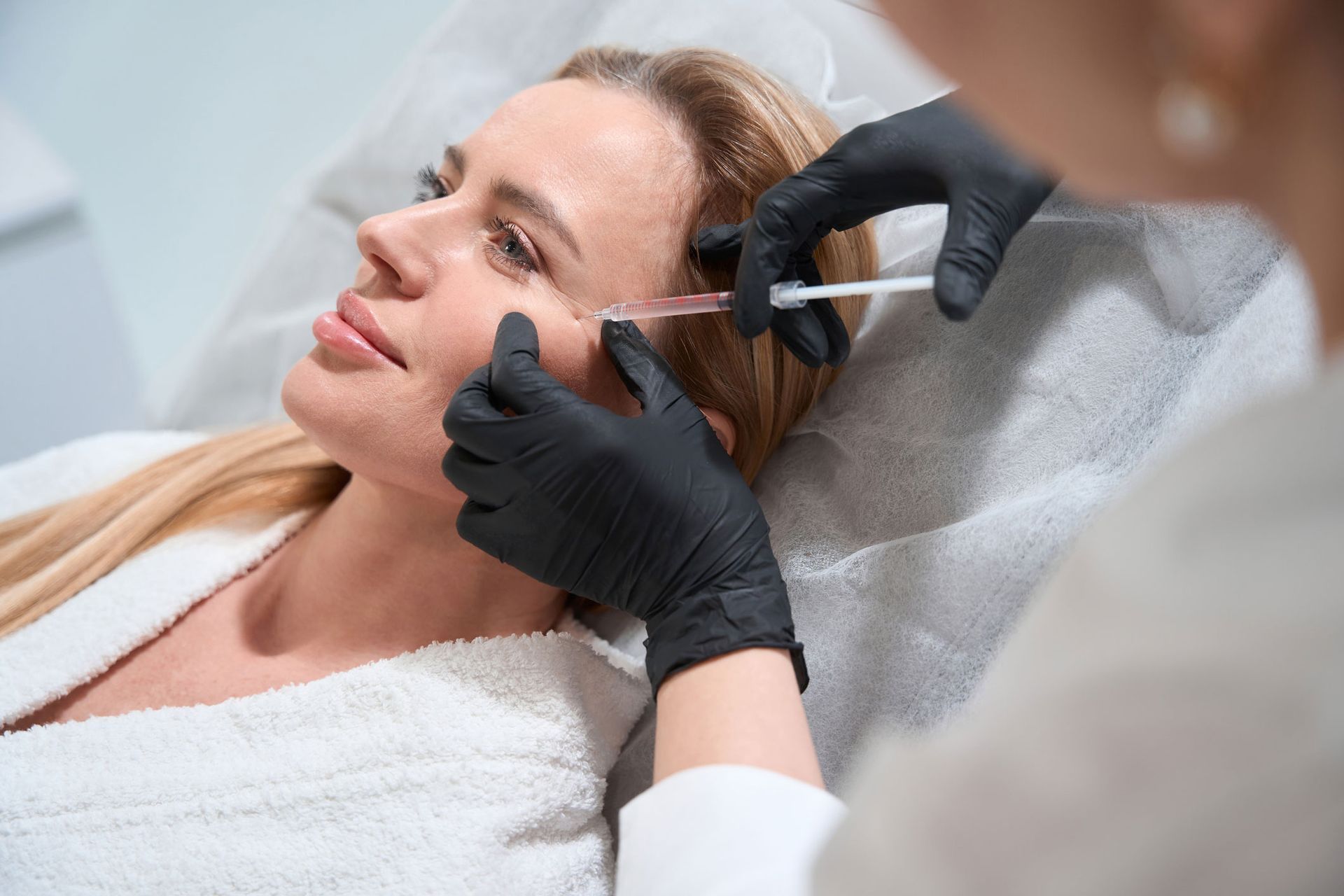 Woman receiving a cosmetic injection near her eye from a person wearing black gloves in a medical setting. Woman receiving a cosmetic injection near her eye from a person wearing black gloves in a medical setting.