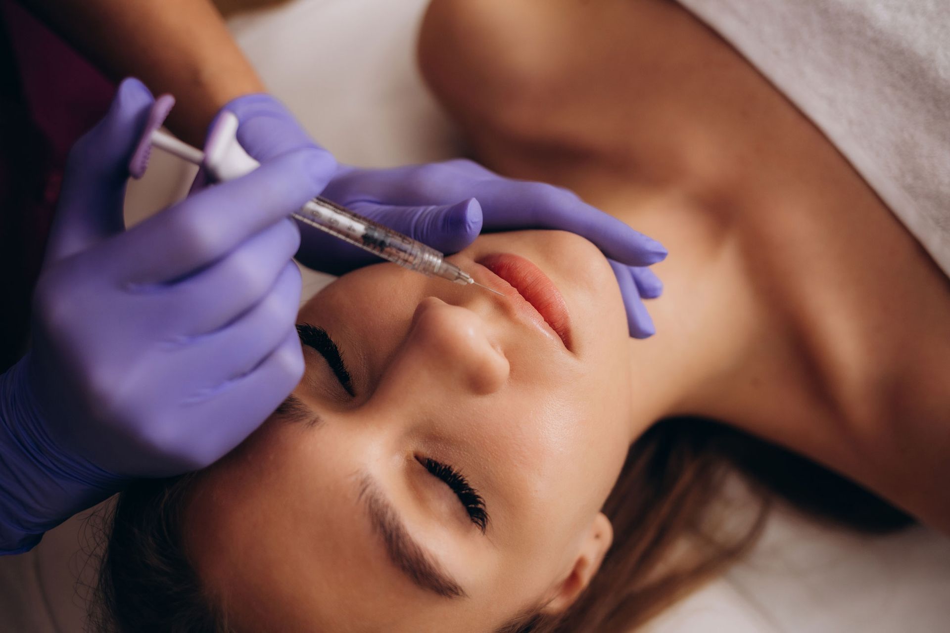 Person receiving lip filler injections. Hands in purple gloves, syringe near lips.