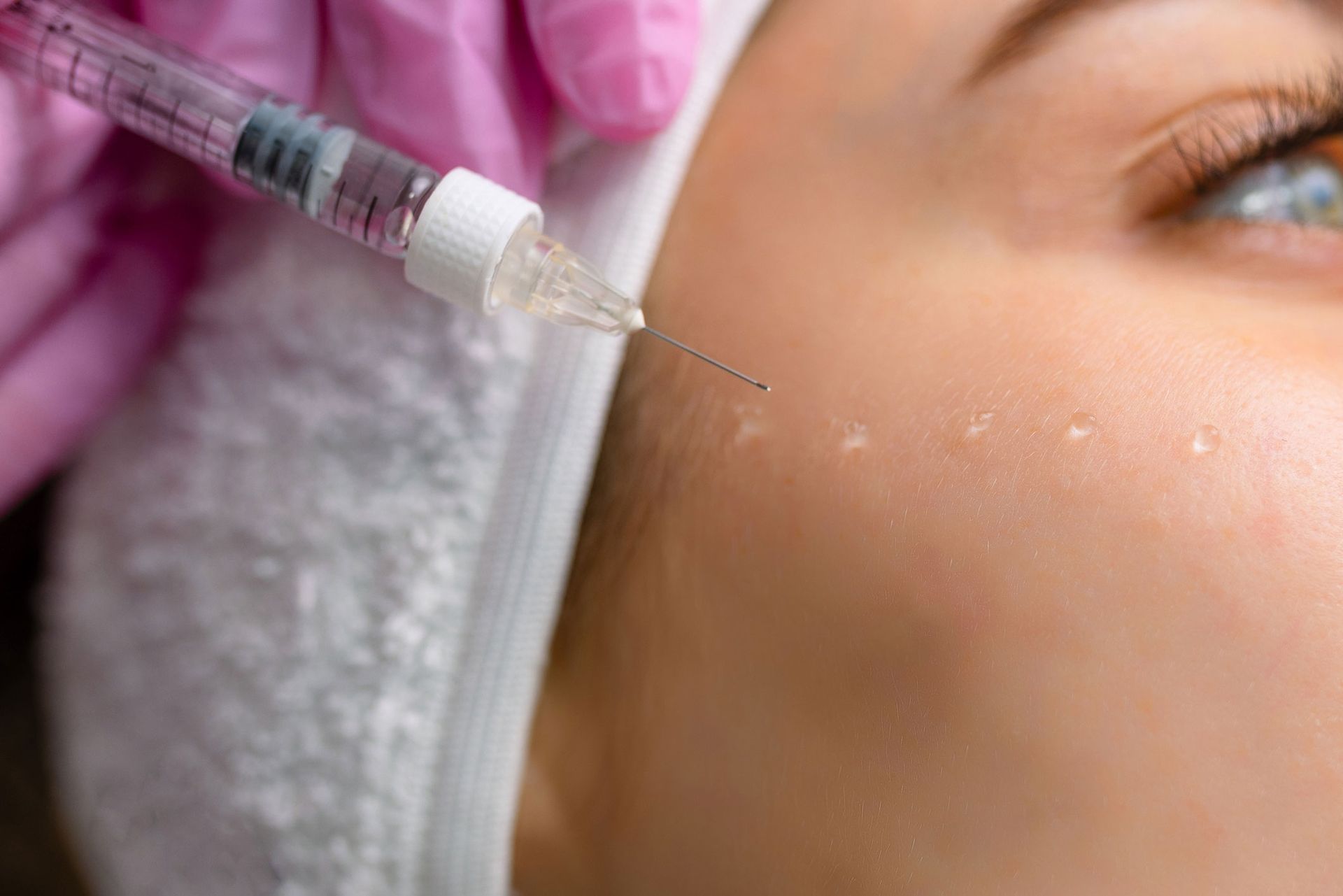 Person receiving a facial injection with a syringe; pink gloves, close-up, white towel, light skin. Person receiving a facial injection with a syringe; pink gloves, close-up, white towel, light skin.