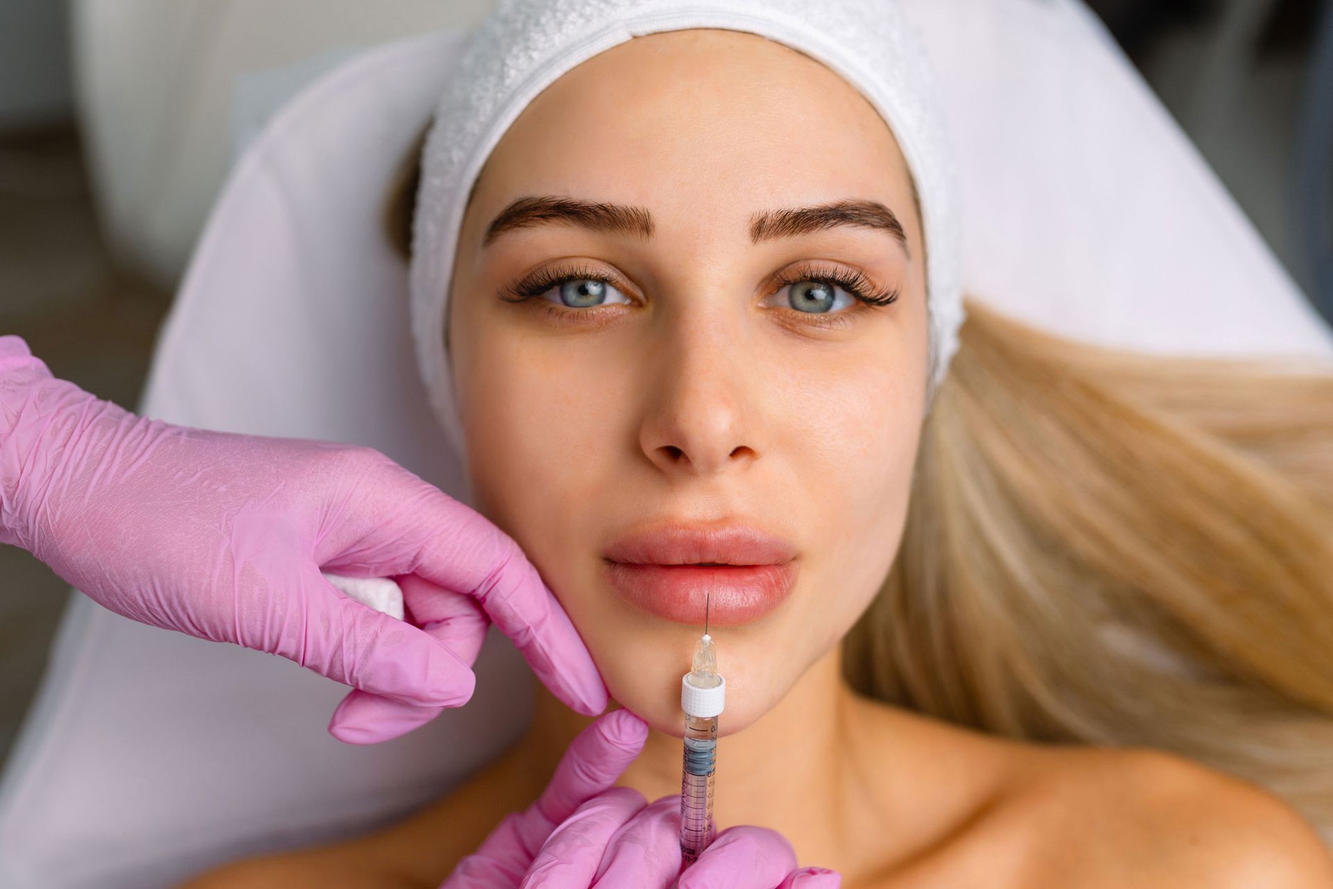 Woman receiving lip filler injections; healthcare professional in pink gloves holds syringe near her lips. Woman receiving lip filler injections; healthcare professional in pink gloves holds syringe near her lips.