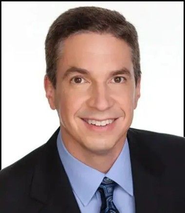 Man in a suit smiling, looking at the camera. Brown hair and eyes, light skin.