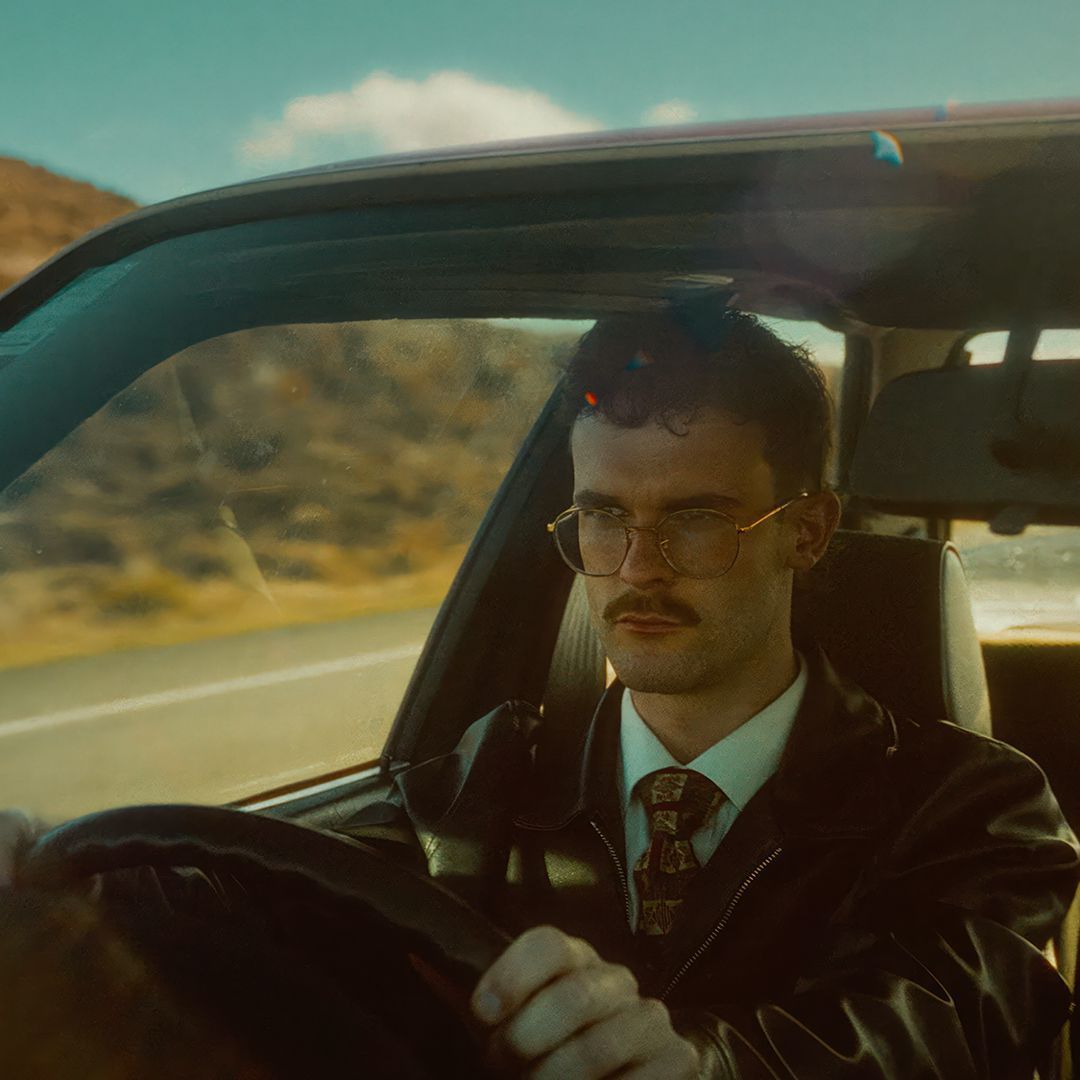 A man with glasses and a mustache is driving a car