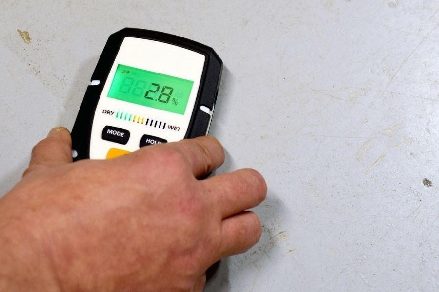 Moisture meter reading elevated levels on a kitchen cabinet floor near a dishwasher, with the kickplate removed to show water staining