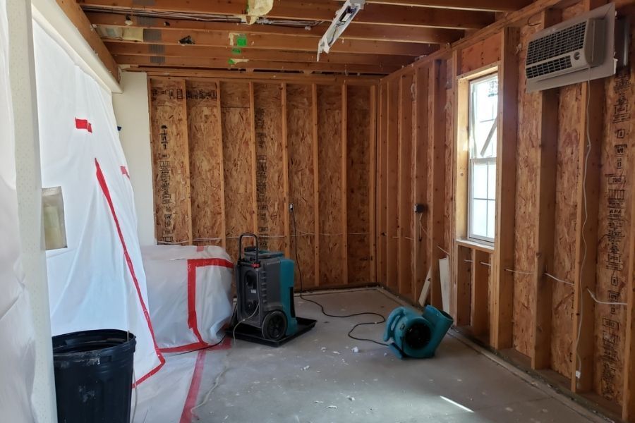 Technician removing wet drywall from behind a bathtub surround, revealing wall studs and insulation, with air movers positioned nearby