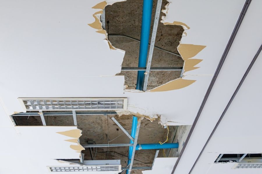 Technician on a ladder inspecting a partially removed ceiling section, revealing wet insulation and darkened wood framing above