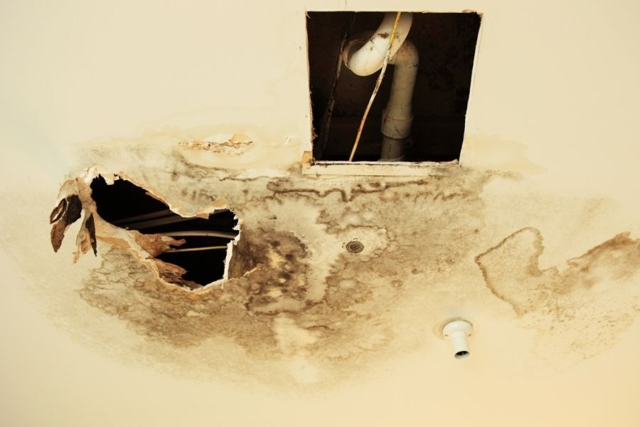 Residential living room ceiling with a spreading brown water stain and slight bulging near the center, light fixture visible nearby