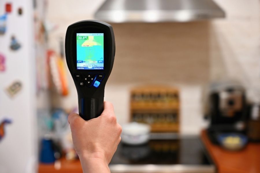 Technician using a thermal imaging camera on a kitchen wall adjacent to a dishwasher, with removed kickplate and extraction hose visible