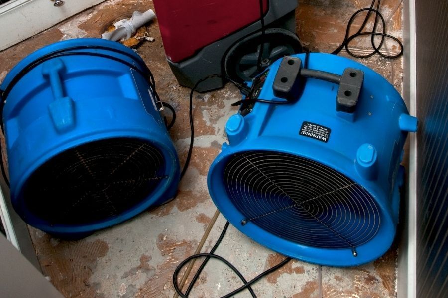 Two technicians deploying air movers and dehumidifiers in a living room with removed baseboards after storm flooding, Central Florida home