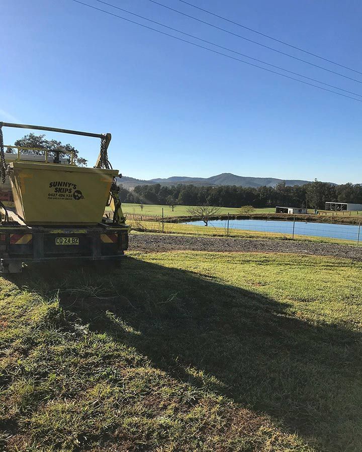 A Yellow Skip Bin Is Parked In A Grassy Field Next To A Body Of Water — Sunny's Skips in Laurieton, NSW