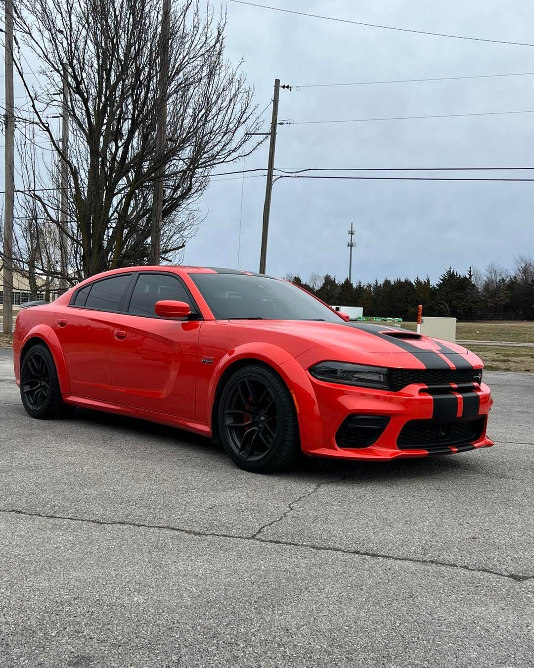 Red Dodge Charger with black racing stripes parked on asphalt, cloudy sky in background.