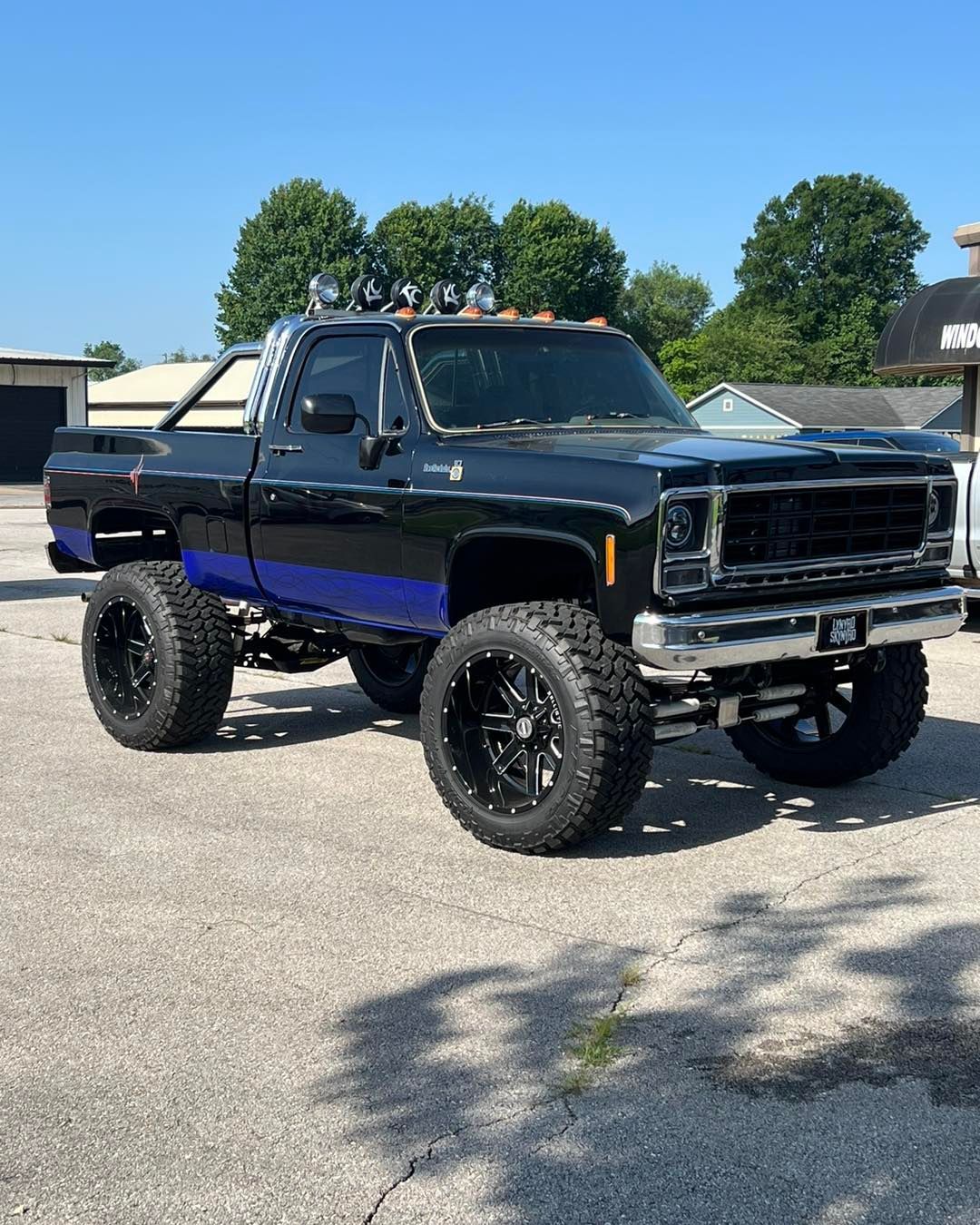 Black lifted truck with blue stripe, large black wheels, and off-road lights, parked on pavement.