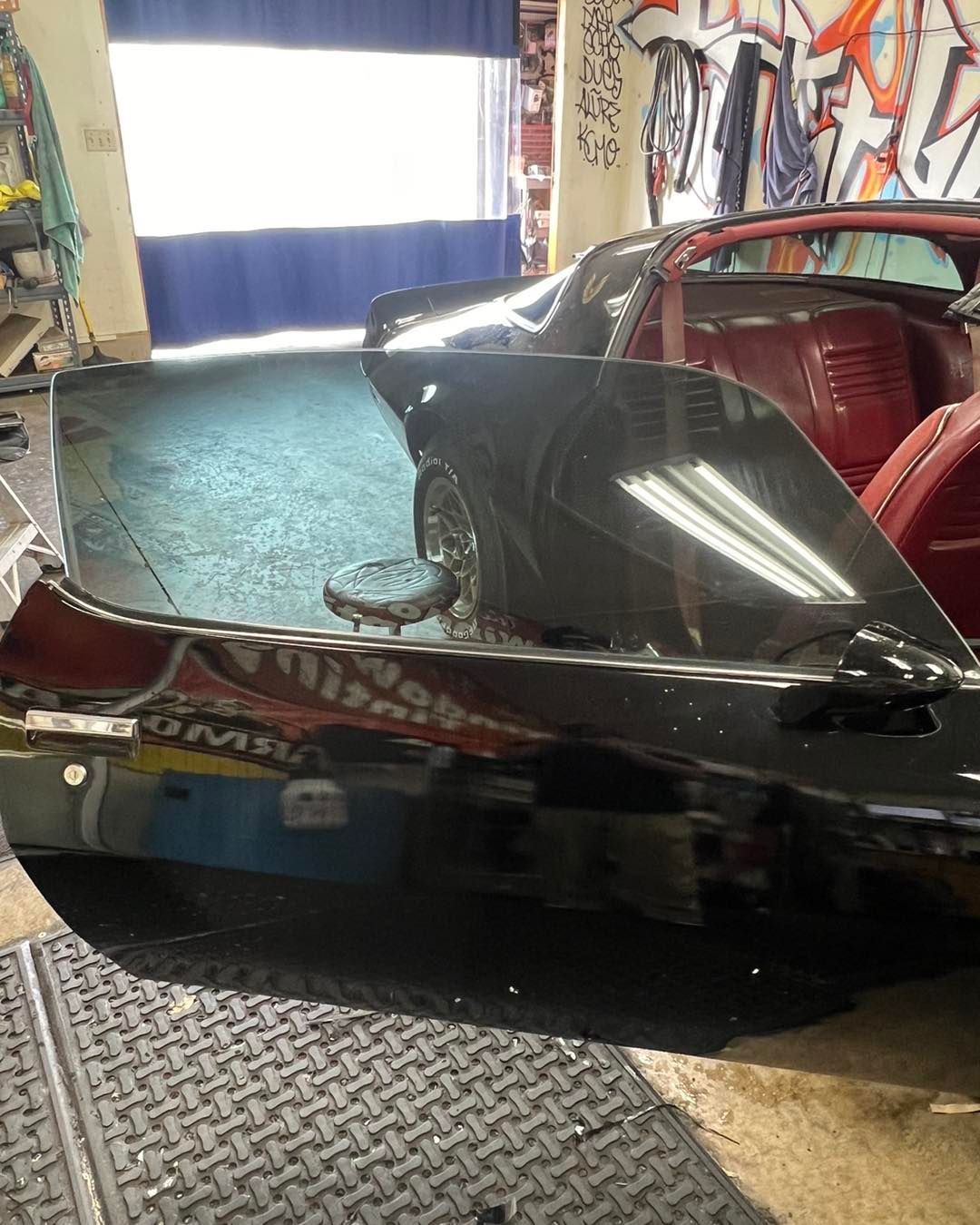 Black car's rear end with a partially open door, interior visible. Shiny paint reflects overhead lights; indoors.