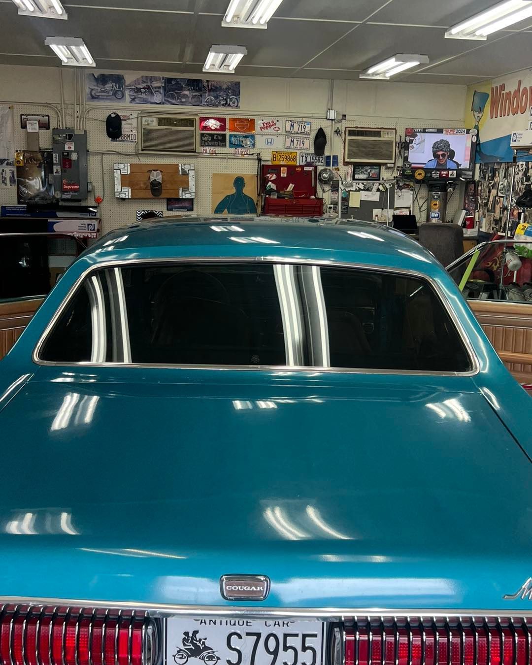 Teal classic car inside a garage, license plate reads S7955. Windows reflect garage lights.