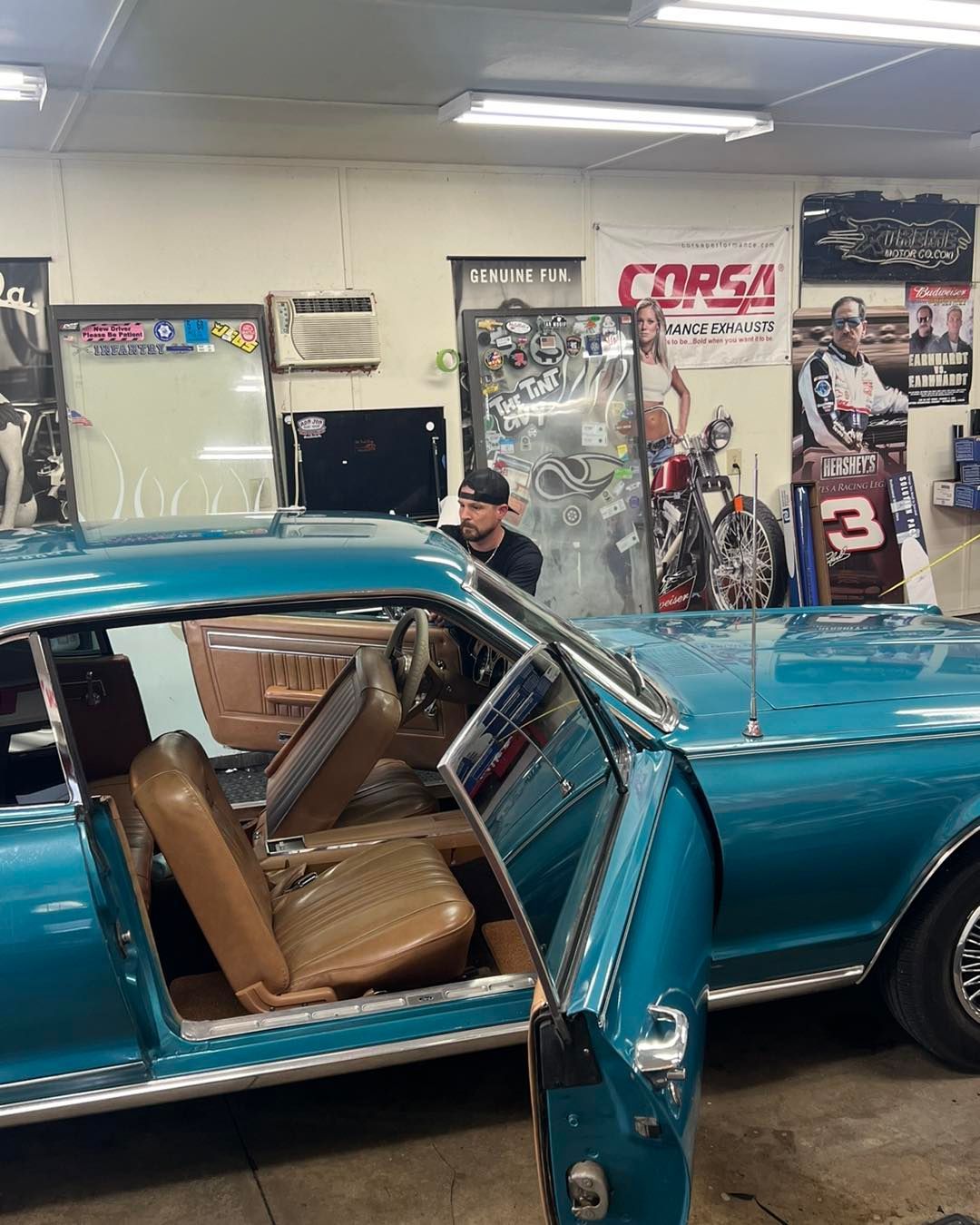 A man working on a turquoise classic car in a garage. Car doors are open, tan interior visible.