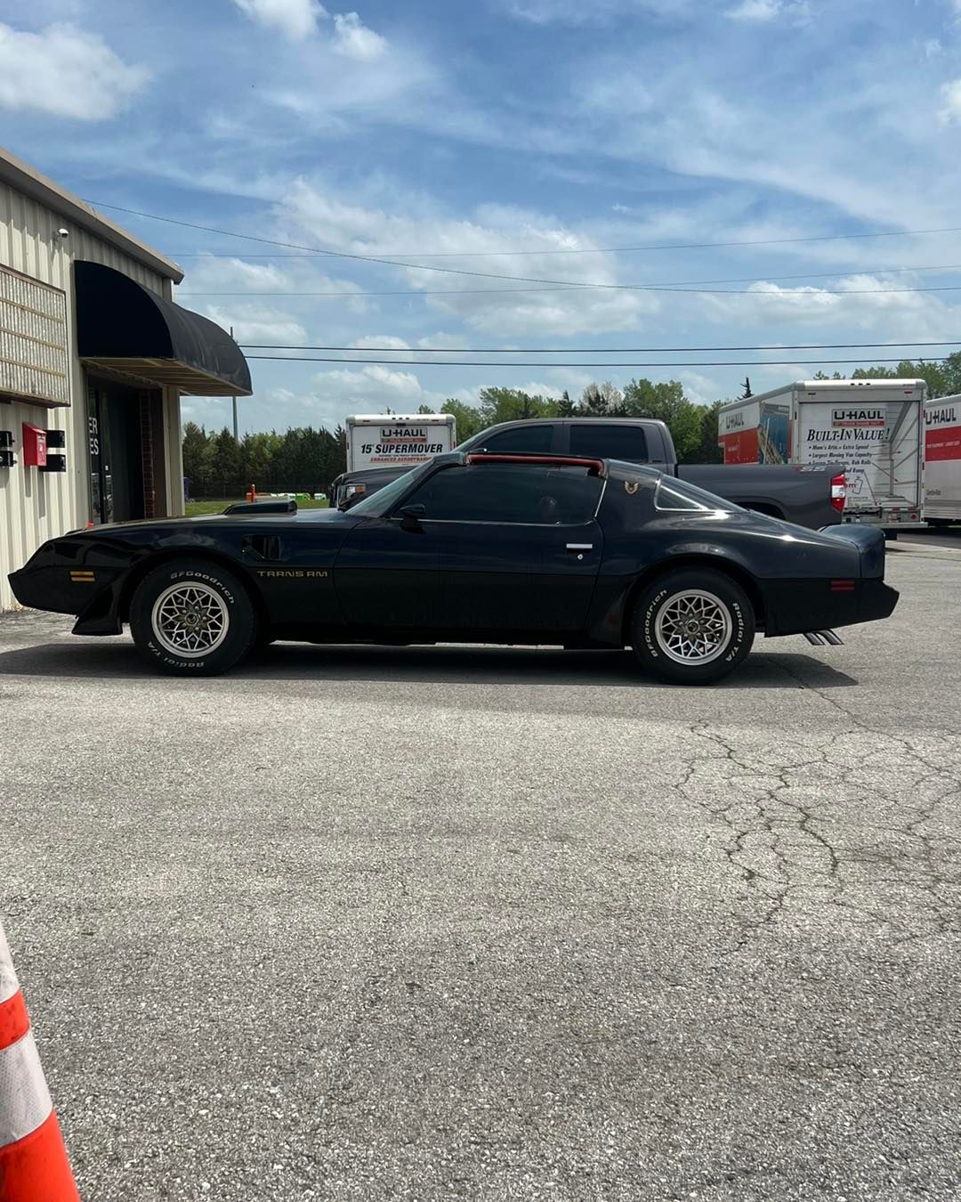 Black Pontiac Firebird parked outside a building, with moving trucks in the background.