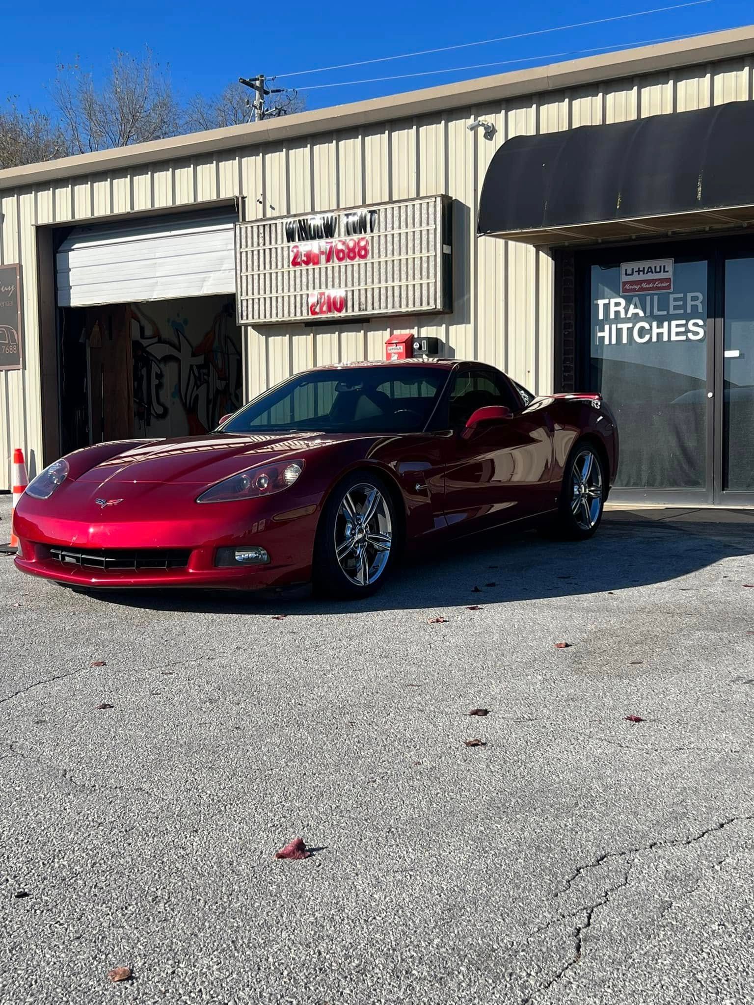 Red Corvette parked in front of a trailer hitch shop on a sunny day.