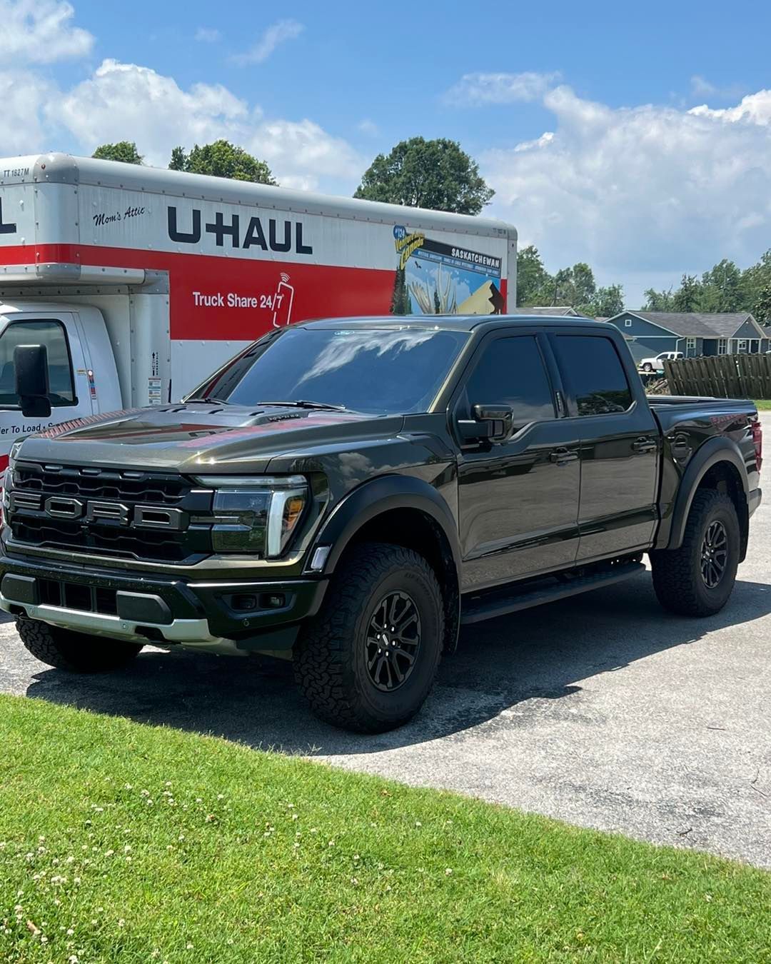 Dark green Ford F-150 Raptor truck parked next to a U-Haul truck on a sunny day.