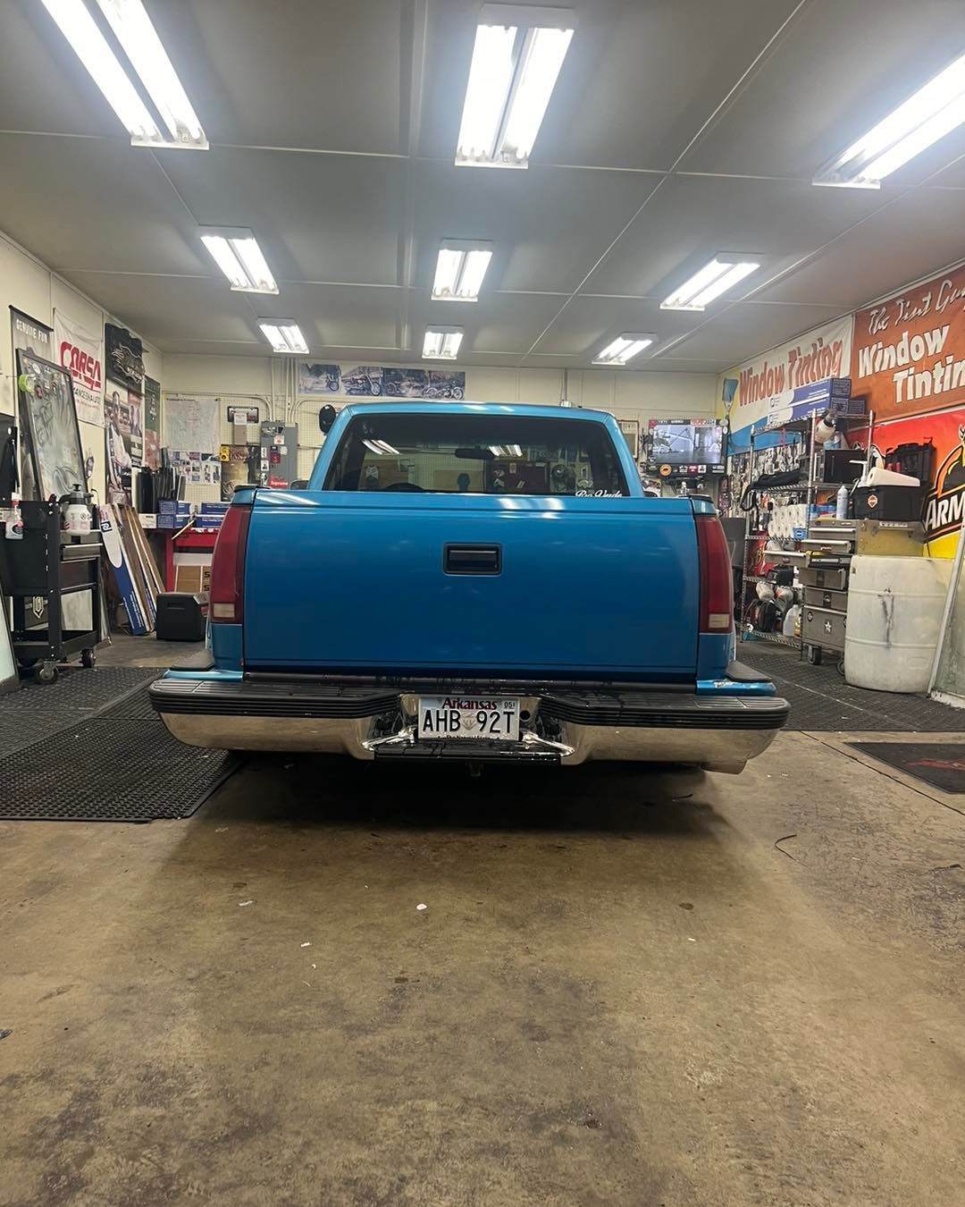 Blue pickup truck's rear view in a garage. Chrome bumper, license plate, and fluorescent lights overhead.