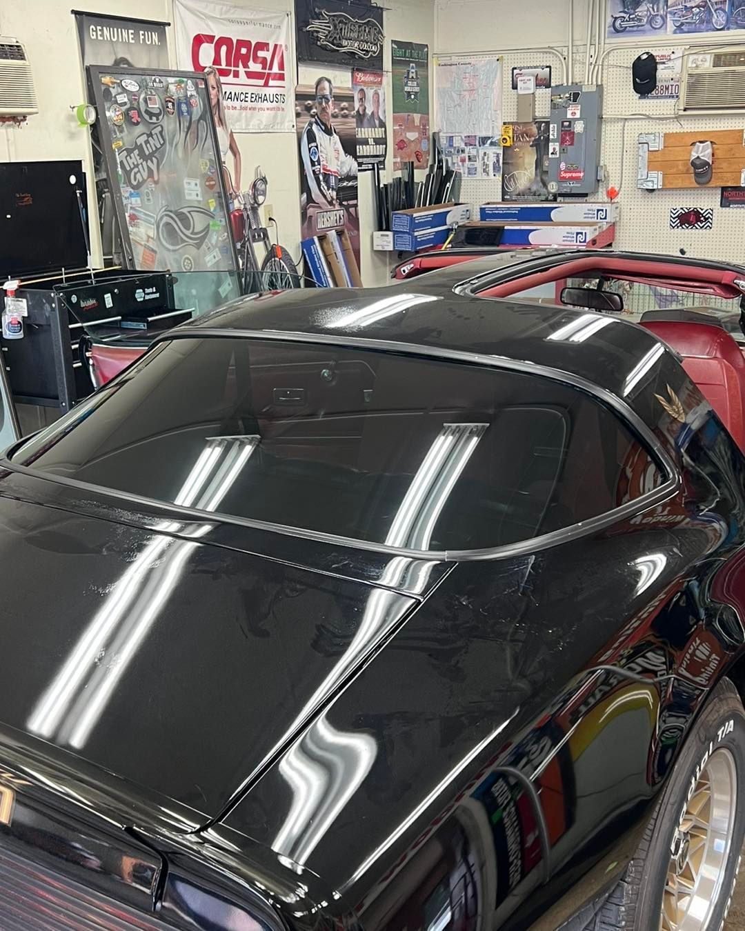 Black sports car with tinted rear window in a garage setting with posters and tools on the wall.