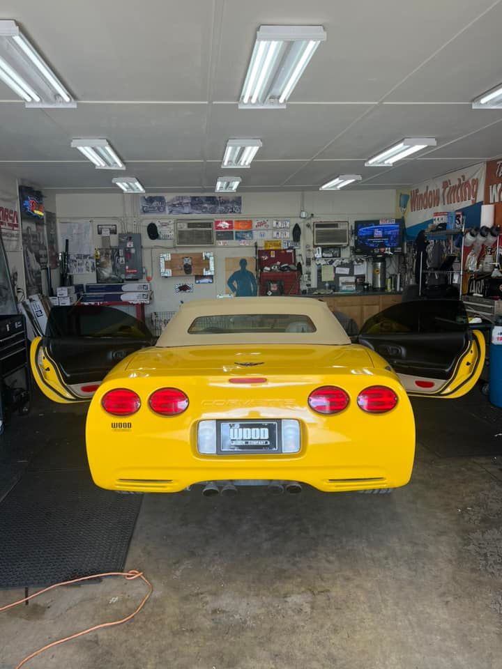 Yellow Corvette convertible with doors open in a garage, under fluorescent lights.