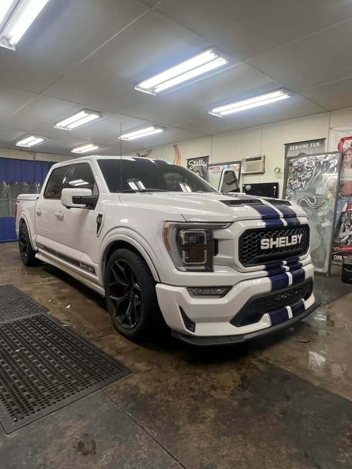 White Ford Shelby F-150 truck with blue racing stripes, parked indoors.