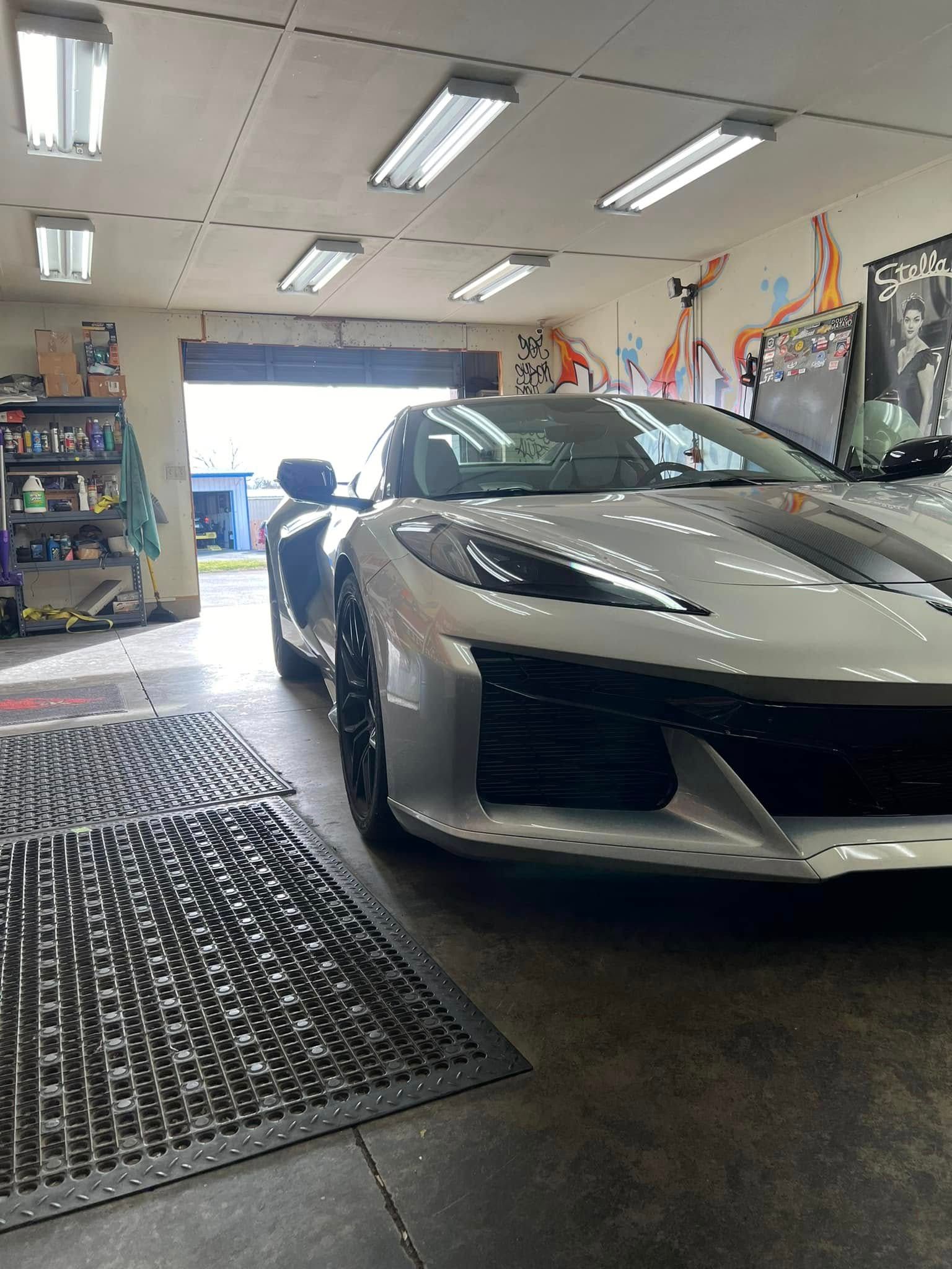 White sports car in a garage with open door, black mat, and graffiti art on the walls.