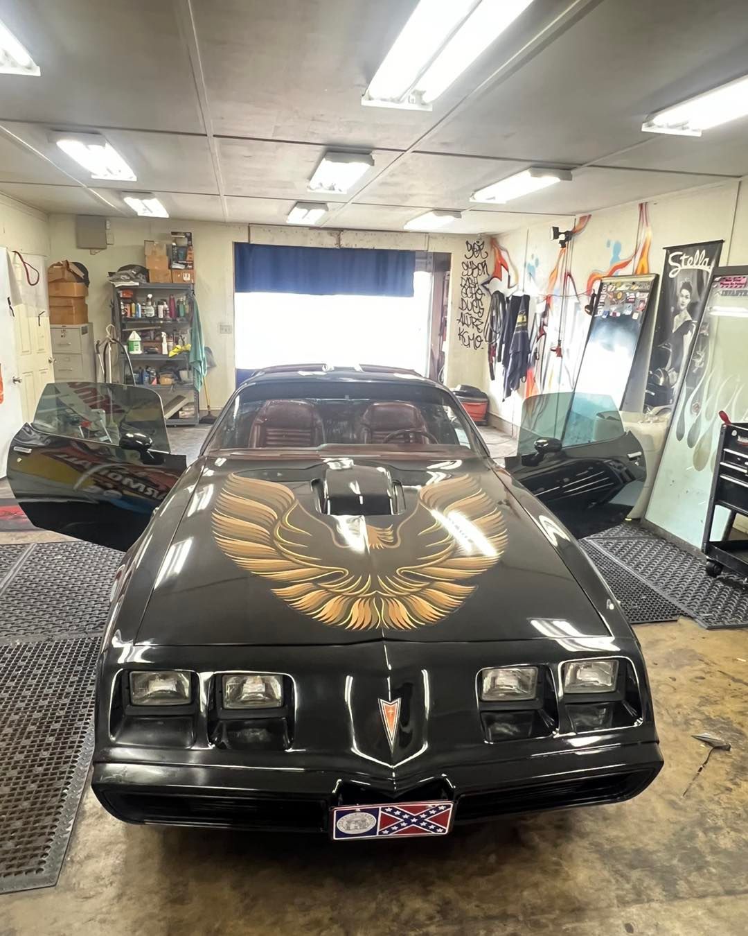 Black Pontiac Firebird Trans Am with gold hood emblem, doors open, in a garage.
