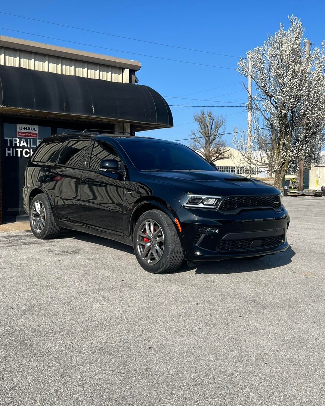 Black Dodge Durango SUV parked in front of a building with a trailer hitch sign, sunny day.