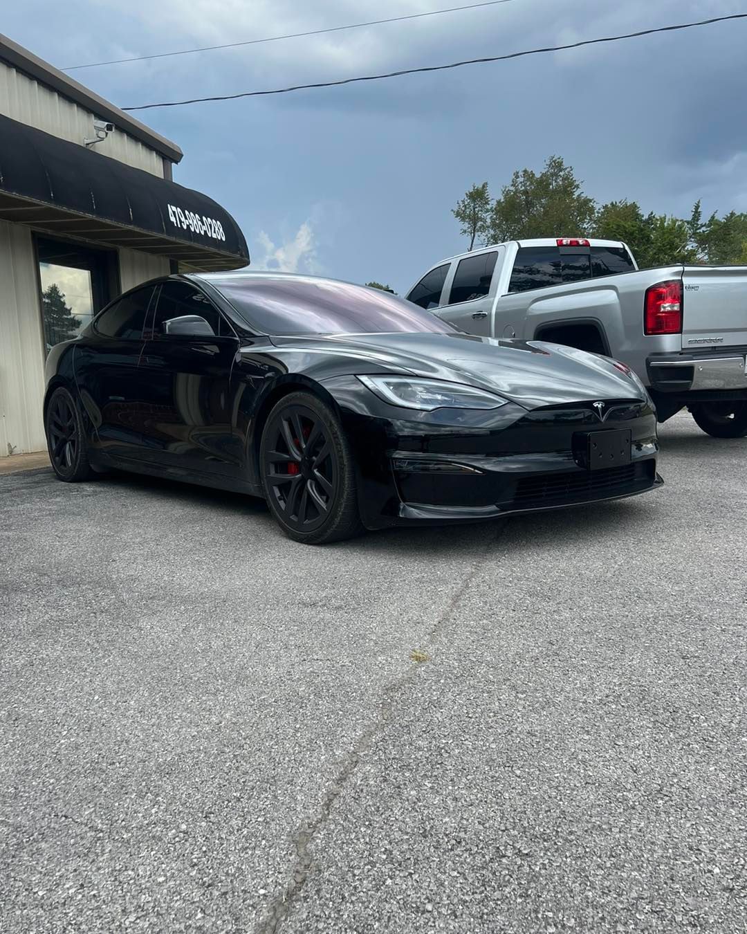 Black Tesla car parked in front of a building with a silver truck beside it, gray pavement, cloudy sky.