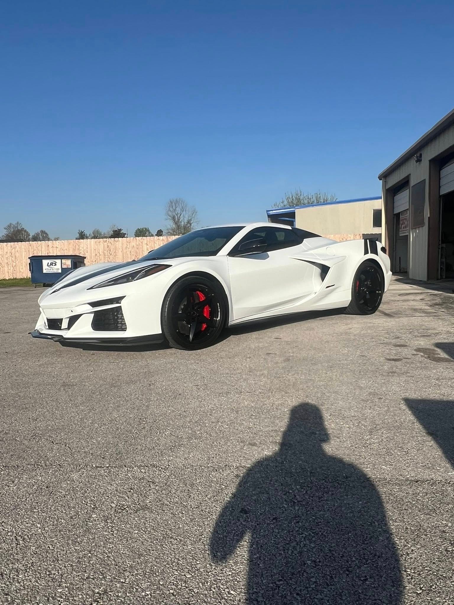 White sports car with black racing stripes parked on gravel; shadow of a person.
