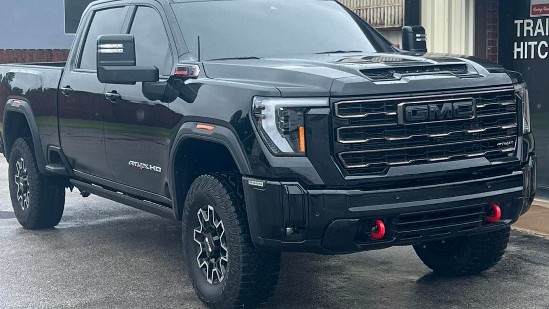 Black GMC Sierra pickup truck, AT4 model, parked in front of a building, gray wheels, red tow hooks.