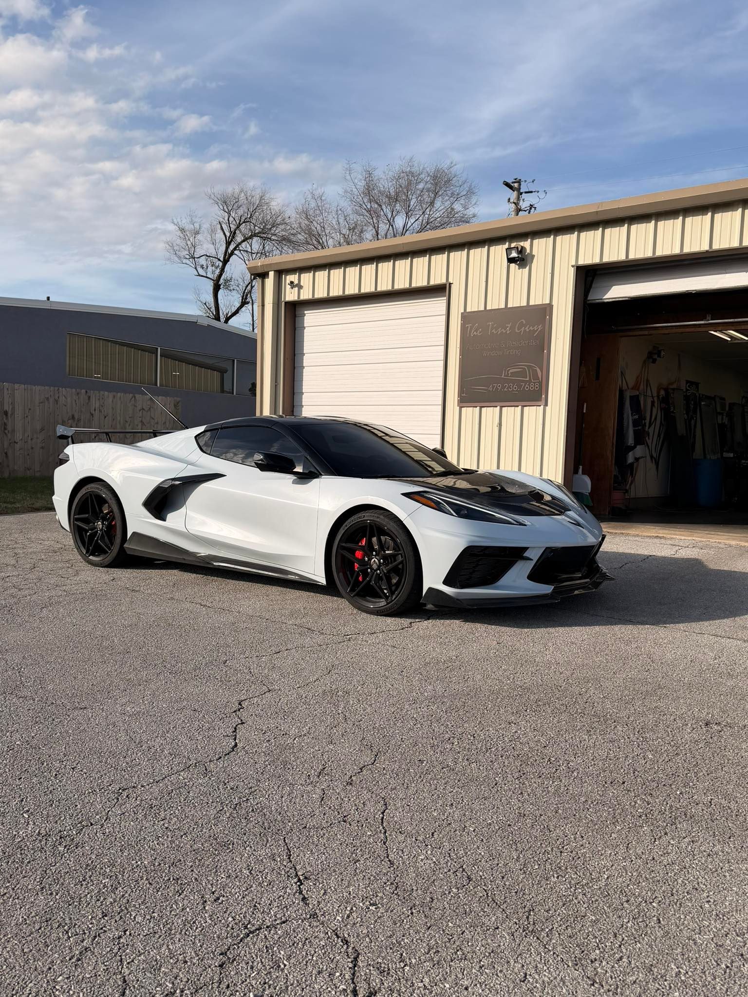 White sports car parked in front of a garage, with a gravel driveway and blue sky.