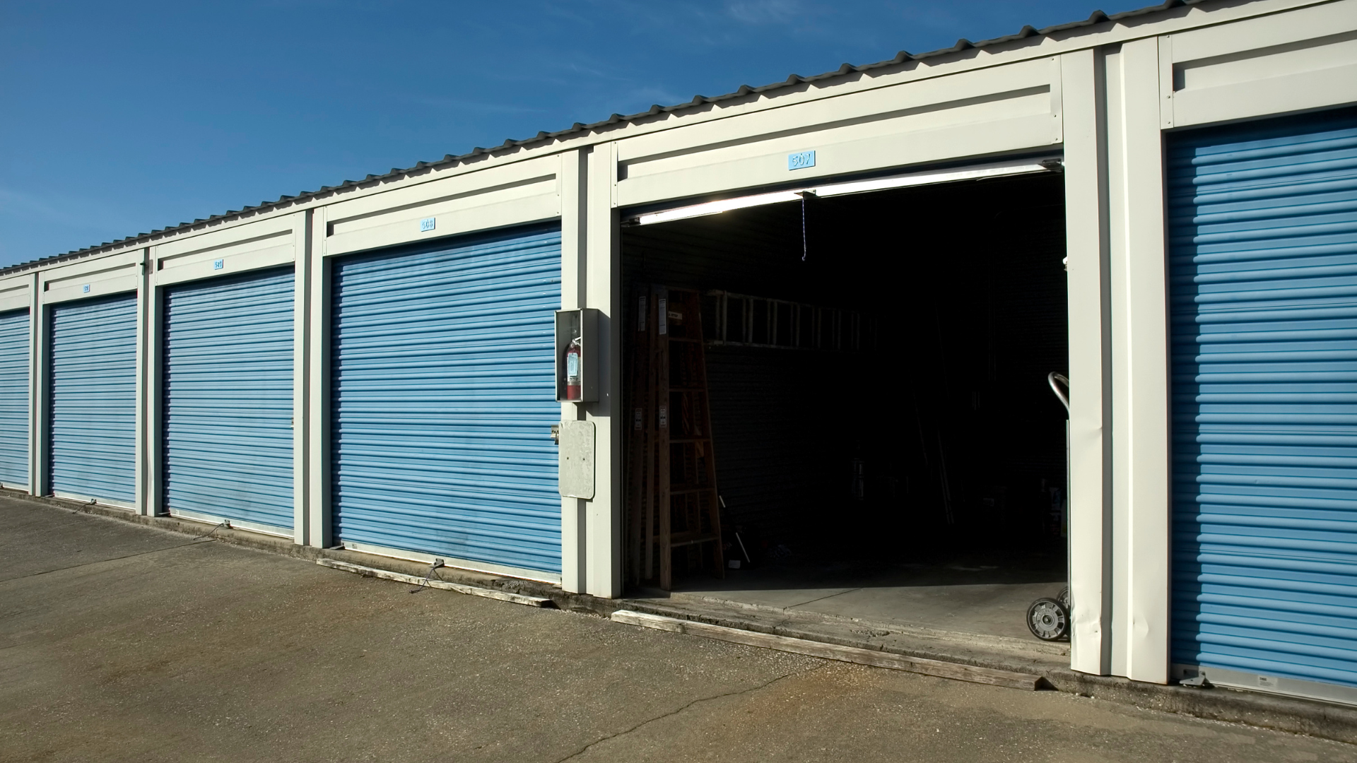 Storage units with blue rolling doors; one open, revealing interior. Concrete and blue sky.