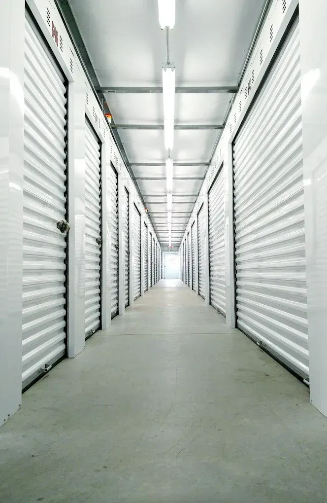 Interior of a storage unit facility with white doors lining a long hallway, lit by overhead lights.